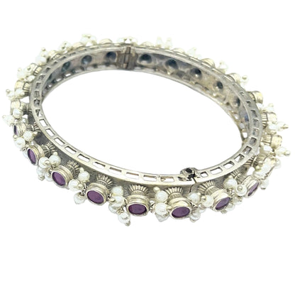 A pair of silver-toned huggie bangles decorated with rows of small clear stones and purple oval gemstones, displayed on a light blue-white background.